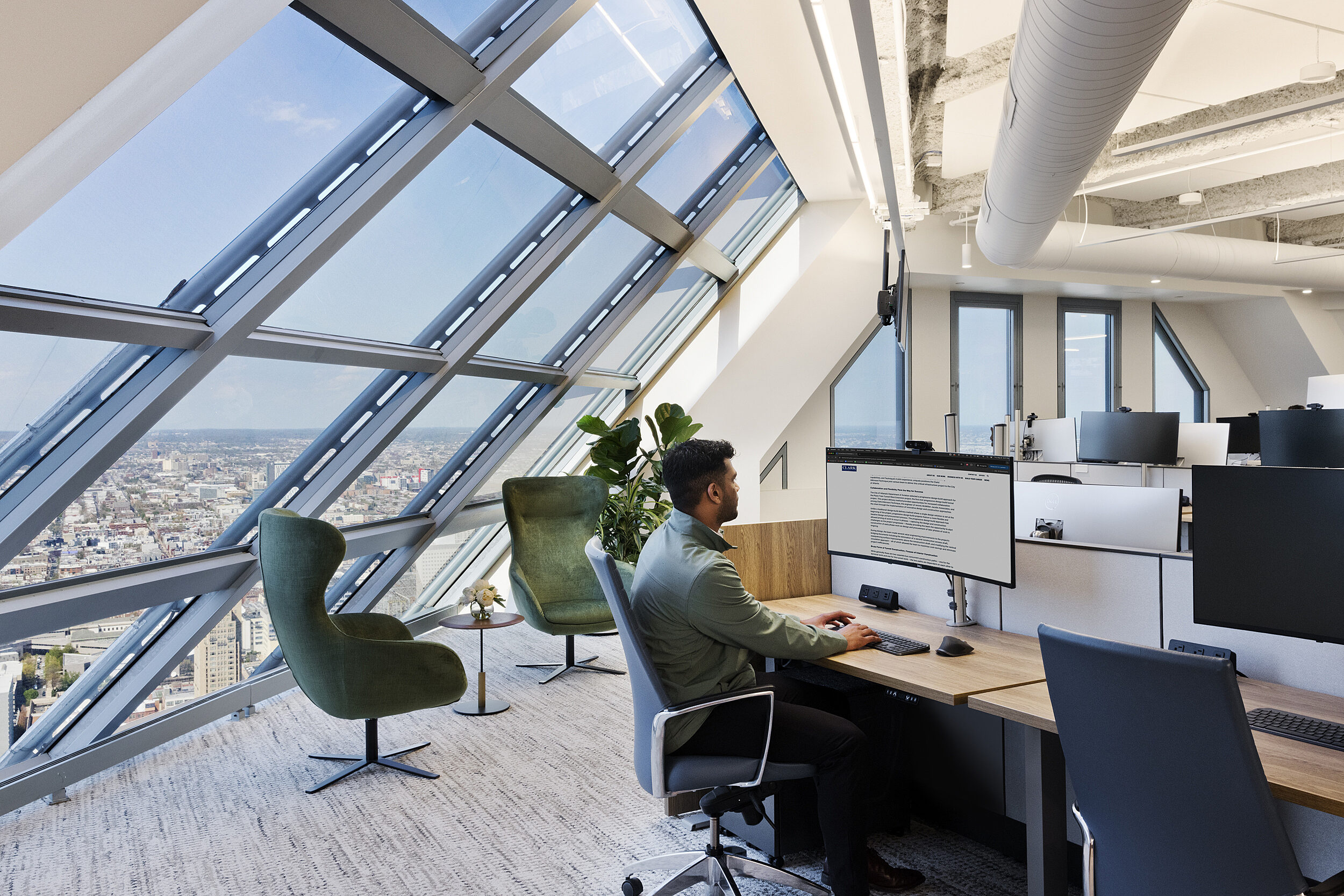 Open office with wood workstations, ergonomic task chairs, dual monitors, and two green lounge chairs by angled windows overlooking the city.