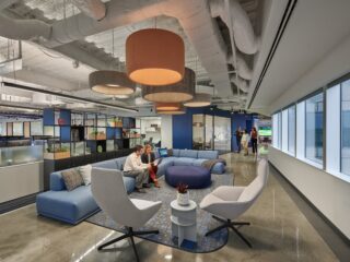 Office lounge with large blue sectional sofa, swivel lounge chairs, round ottoman, small side tables, open shelving, and overhead drum pendant lights.