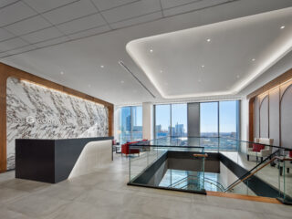 Lobby with a marble reception wall, black reception desk, glass stair railing, and lounge seating with red chairs overlooking city views.