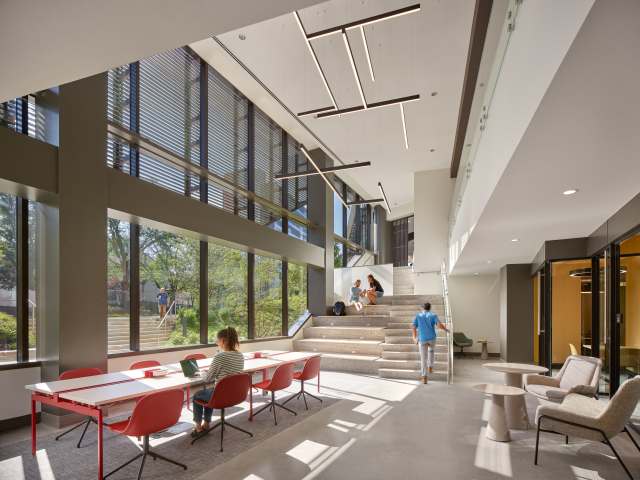 Atrium workspace at university with long white tables and red chairs, seating steps, lounge chairs with small tables, and large floor-to-ceiling windows.