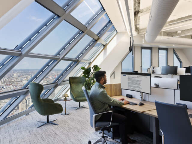 Open office with wood workstations, ergonomic task chairs, dual monitors, and two green lounge chairs by angled windows overlooking the city.