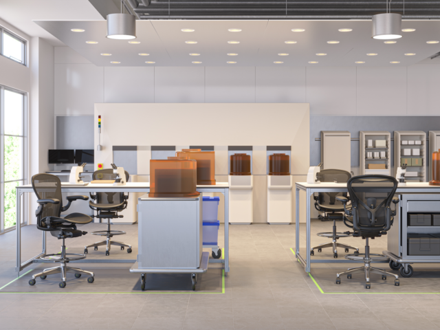 Tech lab with modular workbenches, mesh task chairs, rolling storage carts, and covered equipment units arranged in rows with shelving in the background.