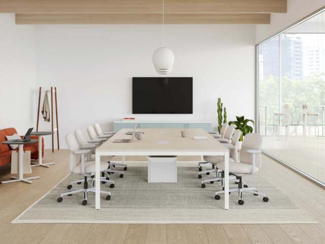 Conference room with a large light wood table, white task chairs, wall-mounted screen, sideboard cabinet, and glass wall with views of the outdoors.